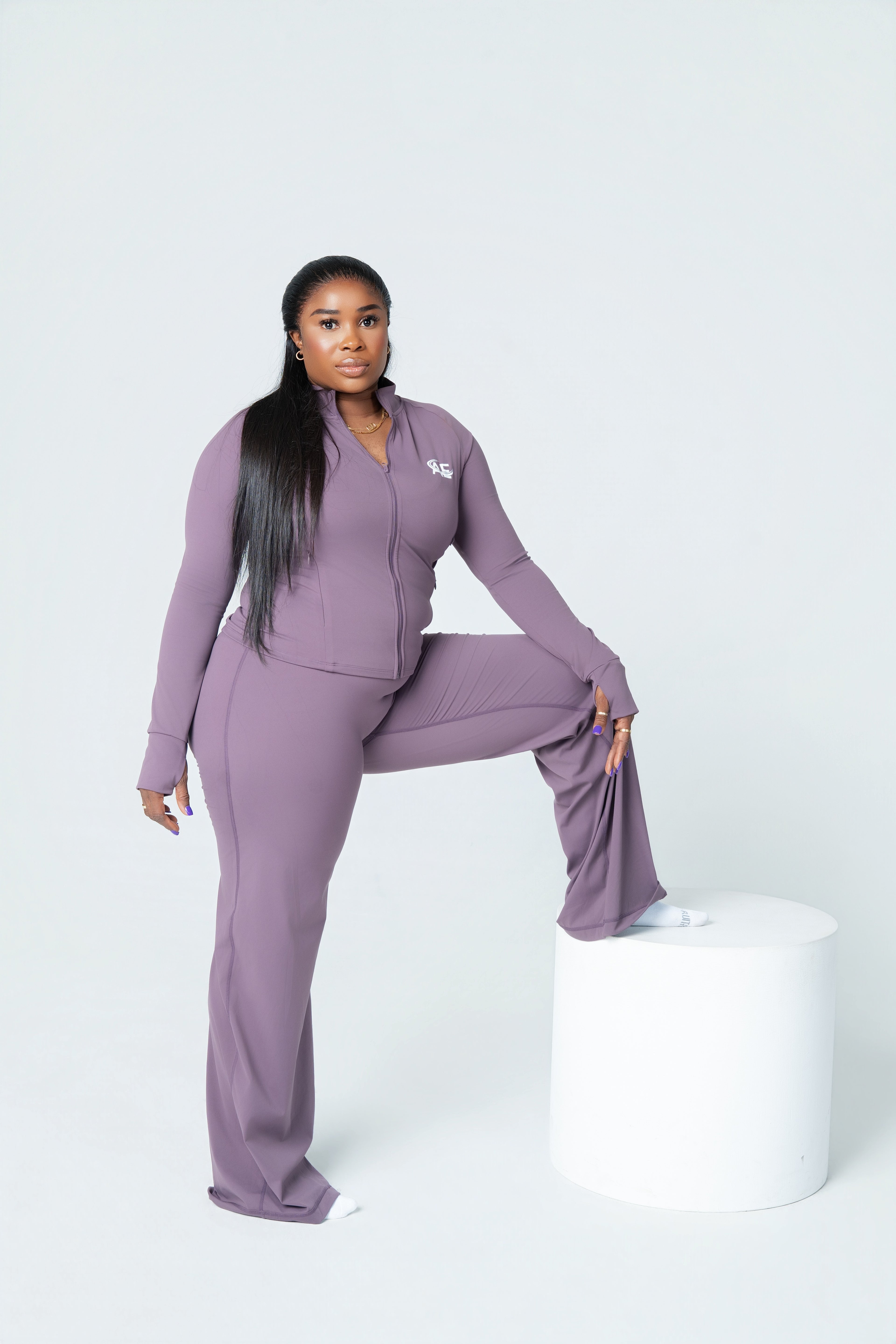 A woman modeling a matching mauve purple tracksuit consisting of a zip-up long-sleeve top and wide-leg pants, posing on a white cylindrical pedestal against a light gray studio background.